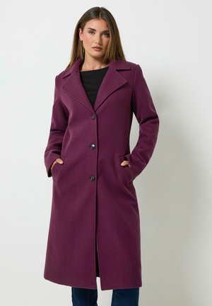 Femme portant un long manteau prune boutonné avec de larges revers, une chemise noire et les mains dans les poches du manteau, debout devant un fond uni.