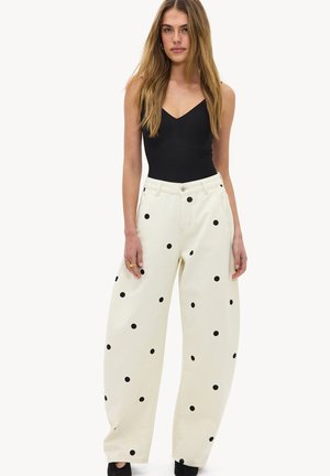 Femme portant un haut noir sans manches et un pantalon blanc à jambes larges taille haute avec des pois noirs, debout devant un fond blanc.