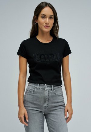 Mujer con una camiseta negra que tiene el texto "SALSA" y unos pantalones vaqueros gris de talle alto, de pie contra un fondo gris claro liso.