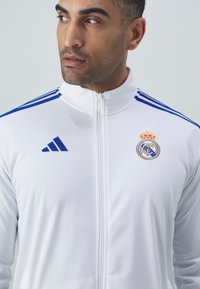 Hvit polyester zip-up jakke med blå detaljer, Adidas-logo og brodert Real Madrid-merke på venstre bryst. Glatt tekstur, atletisk passform.