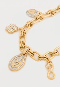 Βραχιόλι με χρυσή αλυσίδα που διαθέτει διάφορα φυλαχτά: ένα τριφύλλι με πολύτιμους λίθους, ένα σχήμα καρδιάς, ένα σύμβολο απει infinito και ένα οβάλ μενταγιόν με λουλουδένιο σχέδιο.