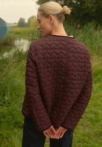Burgundi spalvos siuvinėta striukė su tekstūriniu širdies raštu, turinti laisvą kirpimą ir platėjančias rankoves, dėvima virš tamsių kelnių, lauke ant žolės.