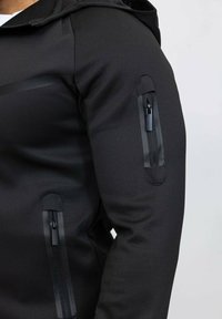 Veste zippée noire avec un design ajusté, featuring un tissu extensible, des accents en panneaux latéraux et des poches zippées sur les manches.