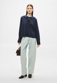 Μπλε σκούρο μπλούζα με μακριά μανίκια, γιακά με βολάν και λεπτομέρεια δέσιμου, σε συνδυασμό με ανοιχτό μπλε παντελόνι φαρδύ και καφέ τσάντα.