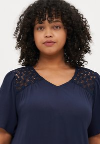 Navy blouse with lace detailing on shoulders, short sleeves, and a V-neckline. Textured fabric with a soft finish.