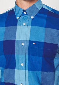 Hombre vestido con una camisa de cuadros azules de botones con botones blancos y un pequeño logotipo en el bolsillo del pecho.