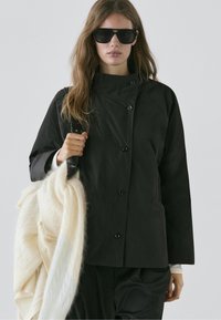 Zwarte jas met knopen en een hoge kraag, voorzien van zijzakken, gedragen over een witte trui, gecombineerd met grote zwarte zonnebrillen.