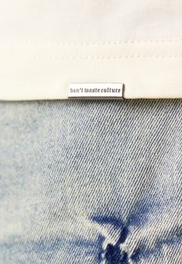 Beige stoffen label genaaid op lichtblauwe denim, met de tekst "verspill geen cultuur" in een vette letters. Tekst is zilverkleurig.