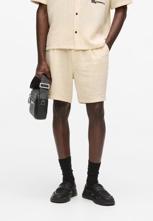 Mann trägt beigefarbene, strukturierte Shorts und Hemd, schwarze Socken und Loafer, hält eine schwarze, rechteckige Ledertasche mit Schulterriemen.