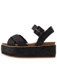 Svart flätad plattformssandal med ett brett vävt rem och justerbart spänne. Med en tjock, strukturerad sula och en tan fotbädd.