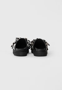 Black sneakers with rubber soles, featuring textured black leather, decorative laces, and metallic accents at the heel.