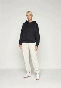 Svart hettegenser med frontlomme, kombinert med kremfargede joggebukser. Fottøyet er hvite sneakers med grå detaljer.