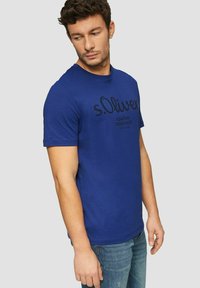 Navy blue cotton t-shirt featuring a round neckline, short sleeves, and black printed logo text on the front. Worn with blue jeans.