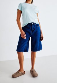 Tekonihkankaiset shortsit, siniset, polvipituiset, joissa on etutaskut ja vyötärökappale. Vaalean sininen lyhythihainen t-paita pienellä vaaleanpunaisella logolla. Harmaat puukengät.