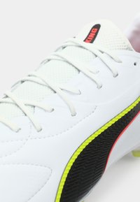 Biała piłkarska korkotrampa z teksturowanym siateczkowym cholewką, zielonymi i czerwonymi akcentami oraz czarnymi detalami po bokach. Z białymi sznurowadłami i kołkami.