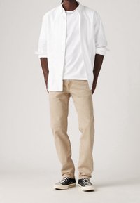 Camisa blanca de botones sobre una camiseta blanca, combinada con jeans beige y zapatillas negras. El conjunto presenta un look casual y en capas.