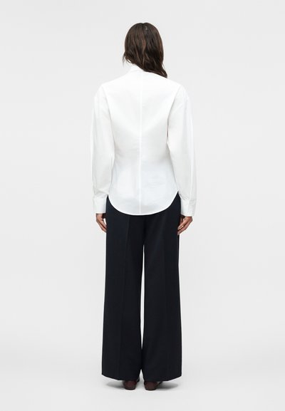 Naine seisab seljaga, kandes keha järgivat valget pikema varrukaga särki ja laiade jalgadega musti pükse ühevärvilisel taustal.