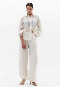 Lätt, genomskinlig vit blus med två framfickor, ihop med vida beige byxor och remmar i guld på klackarna.