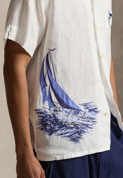 Chemise blanche à manches courtes avec un motif de voilier bleu sur l'eau, portée par une personne à la peau foncée et un pantalon bleu marine.