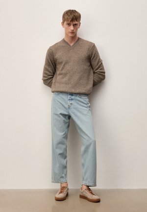 Junger Mann steht vor einer weißen Wand und trägt einen braunen Strickpullover, hellblaue Jeans und beige Turnschuhe mit braunen Schnürsenkeln.