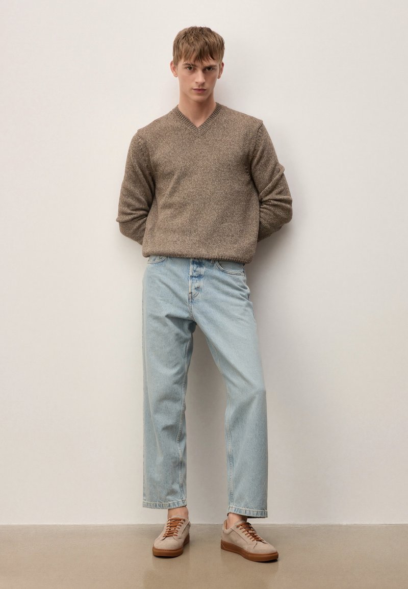 Noor mees seisab valge seina taustal, seljas pruun kudumiga kampsun, helesinised teksad ja beežid tossud, millel on pruunid paelad.