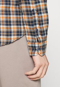 Chemise à carreaux en marine, orange et blanc. Dotée d'un poignet à bouton et d'un tissu texturé avec un motif à carreaux. Main reposant sur une surface beige.