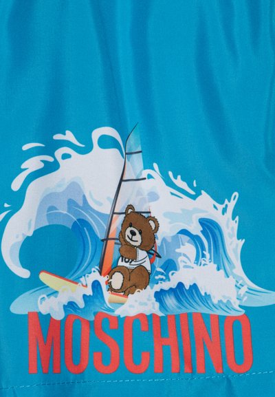 Bañador azul con un personaje de oso sobre una tabla de surf entre olas, con el texto en rojo "MOSCHINO" en la parte inferior. Tejido suave.
