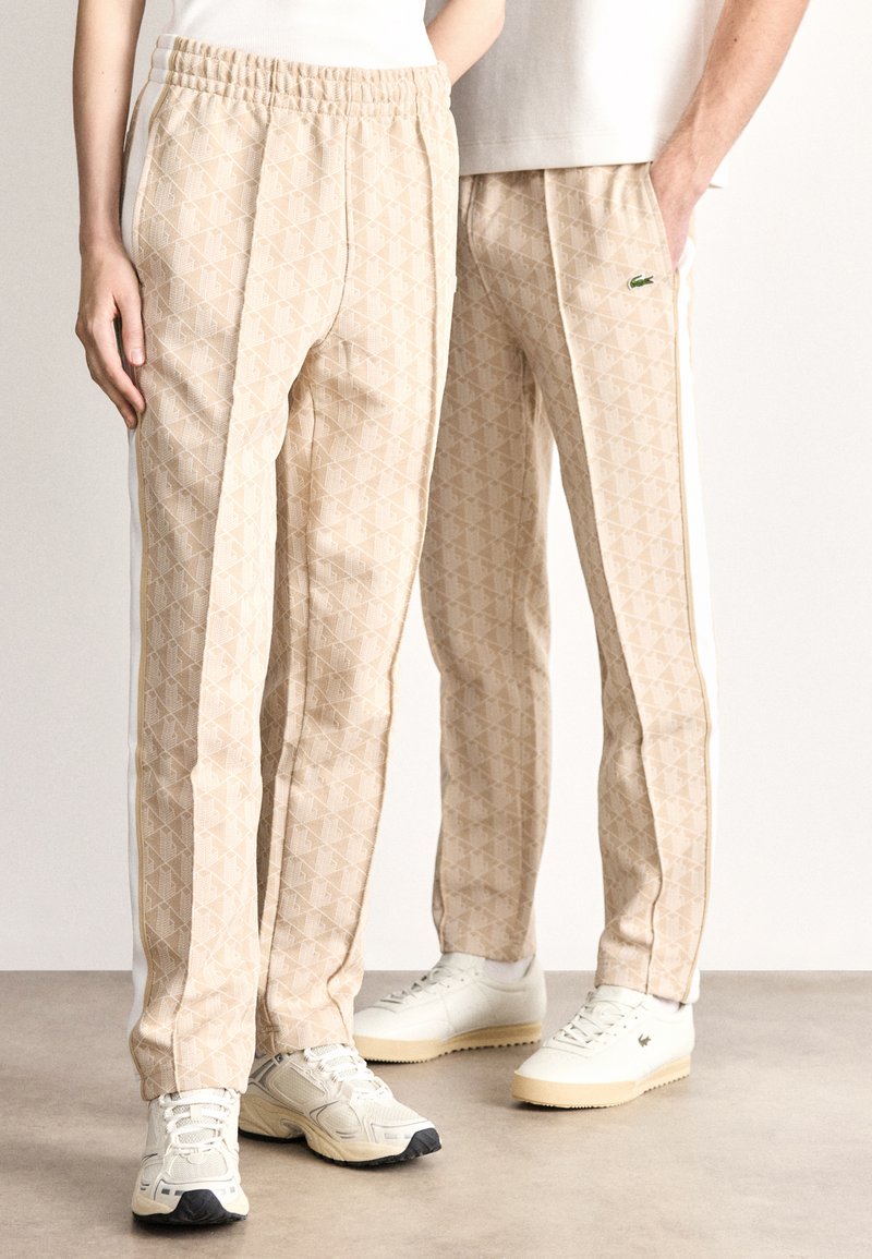 Μπεζ παντελόνια με μοτίβο από απαλό ύφασμα με ελαστική μέση· διαθέτουν λευκές πλαϊνές ρίγες και ένα μικρό πράσινο λογότυπο.