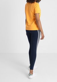 T-shirt orange à manches courtes, leggings navy avec trois rayures blanches, et baskets blanches avec des accents verts, vu de dos.