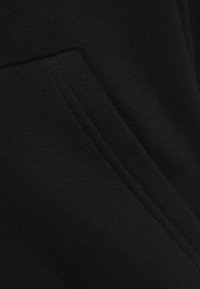 Tissu noir avec une texture lisse et des coutures visibles. Une poche est intégrée dans le design, améliorant la fonctionnalité.