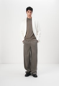 Blazer bianco, maglione marrone e pantaloni larghi grigi chiari, con scarpe nere. Texture lisce, design minimal e palette di colori neutri.