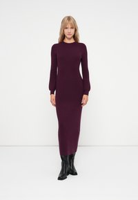 Langes, figurbetontes Kleid aus tiefviolettem Strickstoff, mit Rundhalsausschnitt und langen Ärmeln, kombiniert mit schwarzen knielangen Stiefeln.