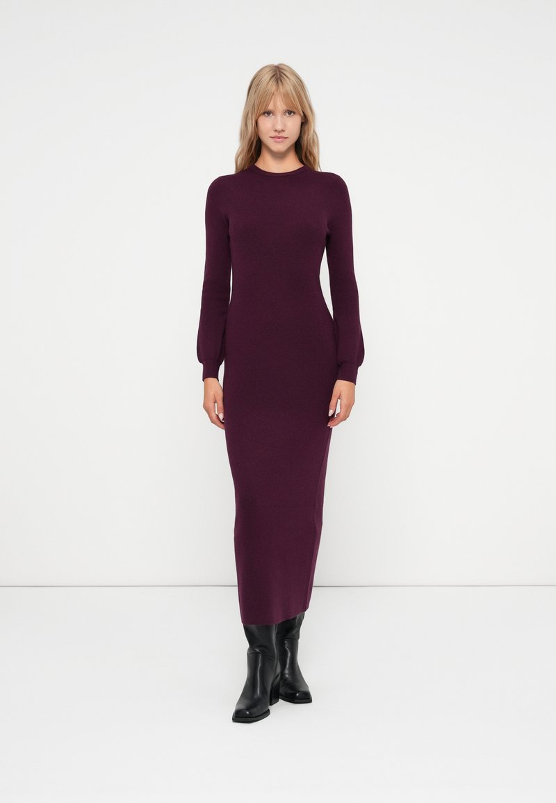 Langes, figurbetontes Kleid aus tiefviolettem Strickstoff, mit Rundhalsausschnitt und langen Ärmeln, kombiniert mit schwarzen knielangen Stiefeln.