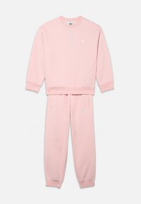 Rozā flīsa džemperis un atbilstošas sporta bikses, abām ar elastīgiem aprocēm, ar nelielu baltu logotipu uz krūtīm un kreisās bikses kājas.