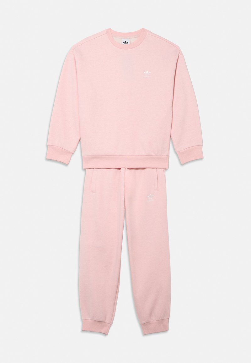 Rozā flīsa džemperis un atbilstošas sporta bikses, abām ar elastīgiem aprocēm, ar nelielu baltu logotipu uz krūtīm un kreisās bikses kājas.