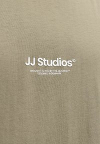 Μπεζ ύφασμα με λευκό κείμενο «JJ Studios» και μικρότερη επιγραφή «Παρουσιάζεται από την ομάδα J&J, σχεδιασμένο στη Δανία» τυπωμένη από κάτω.