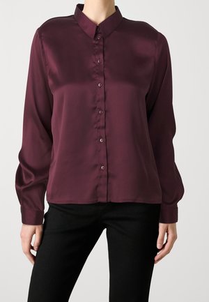 Femme portant un chemisier en satin violet foncé à manches longues et boutonné, ainsi qu’un pantalon noir, debout devant un fond clair uni.