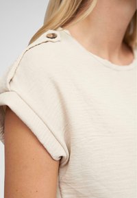 Maglietta beige a girocollo con maniche arrotolate e dettaglio in legno sulla spalla, caratterizzata da un tessuto leggero e testurizzato.