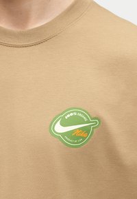 Μπεζ πουκάμισο με πράσινο κυκλικό λογότυπο της Nike που αναγράφει «100% Οργανικό» και «Προϊόν των ΗΠΑ» στο στήθος.