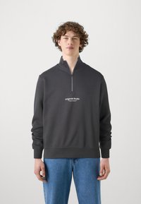 Dunkelgrauer Reißverschluss-Hoodie mit hohem Kragen, ausgestattet mit einem kleinen weißen "originals Studio"-Logo auf der Brust, kombiniert mit hellblauen Jeans.