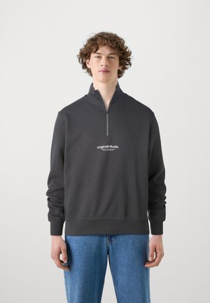 Dark gray zip-up sweatshirt with high collar, featuring a small white "originals Studio" logo on the chest, paired with light blue jeans.