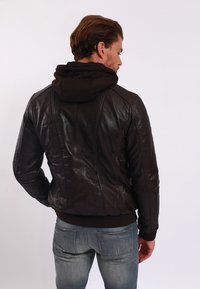 Giubbotto di pelle marrone con cappuccio scuro. Caratterizzato da un design aderente, una texture liscia e cuciture visibili lungo le cuciture posteriori.