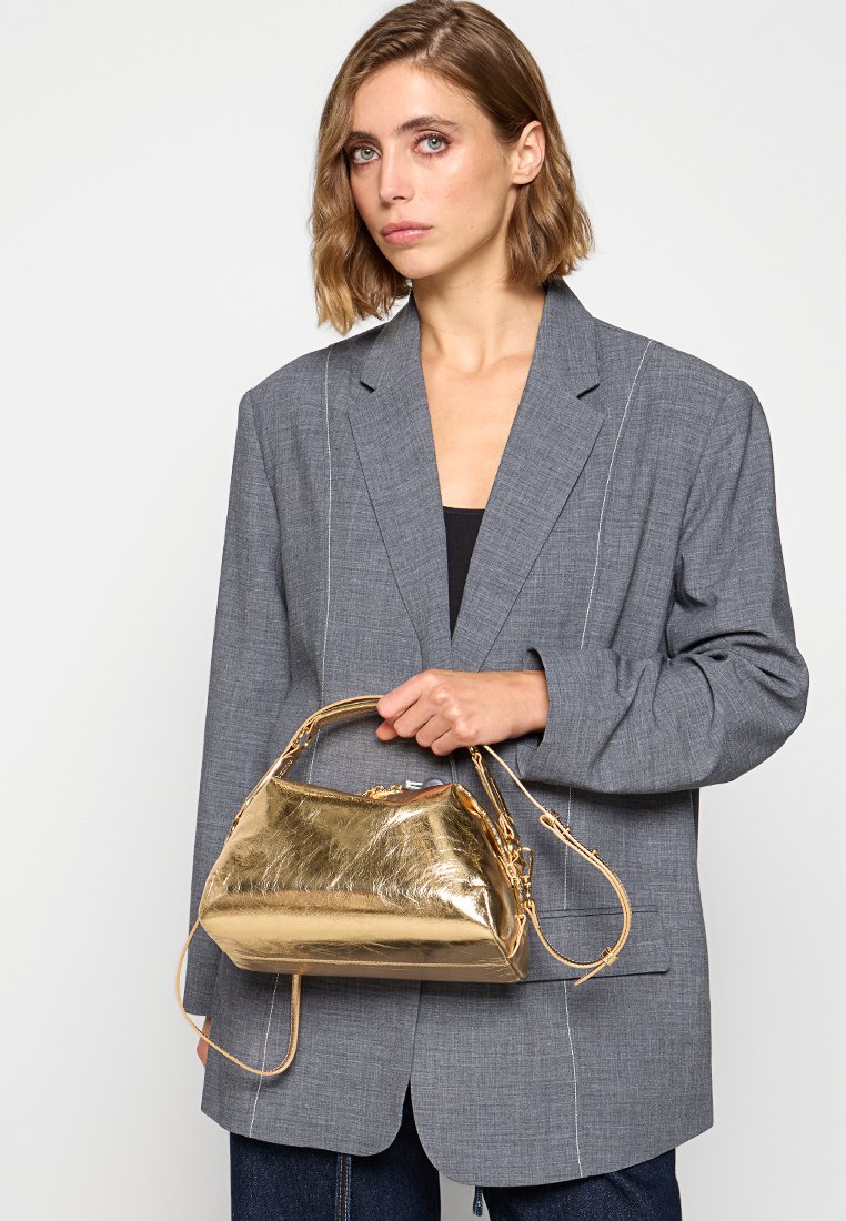 Gouden metallic handtas met een gestructureerde vorm, een gladde textuur, dubbele handvatten en een afneembare schouderriem, vastgehouden door een persoon in een blazer.