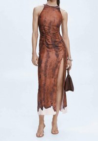 Frau in einem ärmellosen, braunen Batik-Midikleid mit hohem Schlitz, die eine braune Handtasche hält und durchsichtige Riemchensandalen trägt.