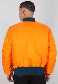 Světlá oranžová péřová bunda s vroubkovaným zeleným límcem a manžetami. Má prošívanou strukturu a elastický spodní lem pro těsný fit.