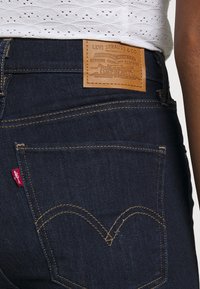 Jeans de mezclilla azul oscuro con una etiqueta de cuero marrón claro que presenta "Levi Strauss & Co." El hilo es de color amarillo, con un distintivo bolsillo trasero de doble arco.