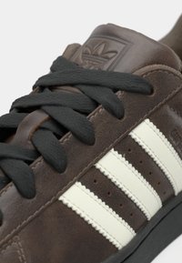 Καφέ δερμάτινο αθλητικό παπούτσι με μαύρα κορδόνια, που διαθέτει τρεις λευκές ρίγες, υφασμένους λεπτομέρειες και λογότυπο στη γλώσσα.