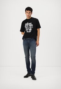 Fekete póló feltűnő fehér grafikával, sötétkék slim fit farmernadrággal és fekete bokacsizmával párosítva. Texturált anyag kontraszt.