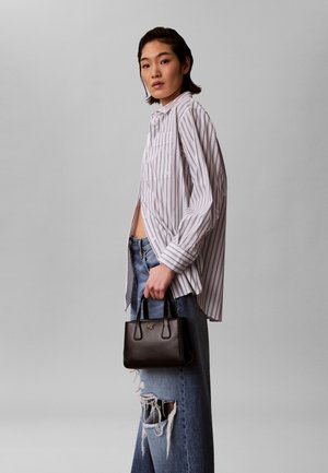 Woman wearing oversized white shirt with vertical stripes and ripped jeans holding a small black handbag against a gray backdrop.