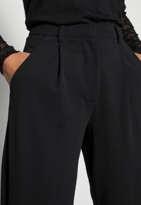 Czarne spodnie z wysokim stanem o dopasowanym kroju, z fałdami, bocznymi kieszeniami i gładkim, lekkim materiałem.